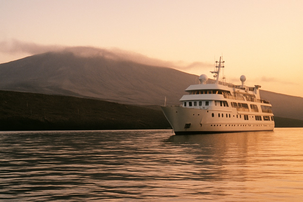 A small expedition ship anchored in a calm bay between volcanic Galapagos islands at sunrise